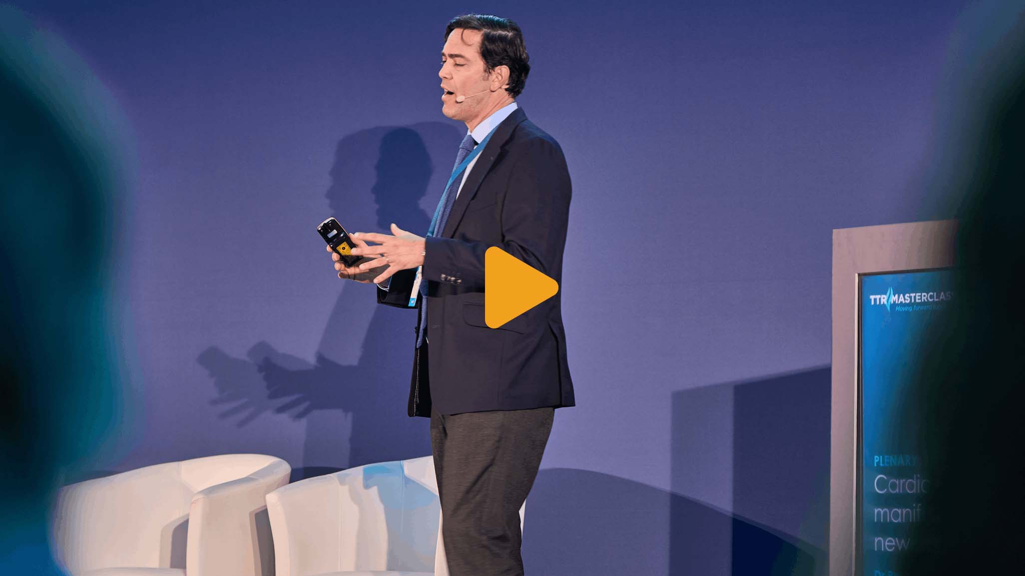 Video thumbnail showing a person in a suit holding a device on stage with a presentation screen in the background