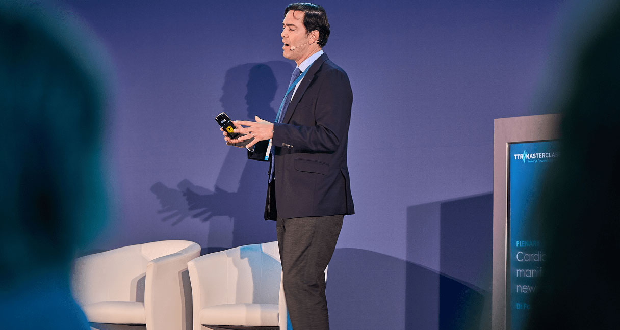 Person in a suit holding a device on stage with a presentation screen in the background